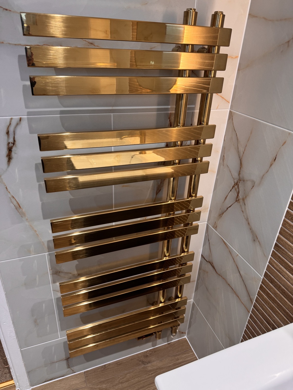 Gold heated towel rail fitted against marble-effect bathroom tiles by SMC
